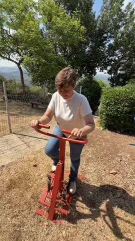 A person with short brown hair, wearing a white t-shirt and blue jeans, is using a red exercise machine outdoors. The machine appears to be a stepper or stair climber, with pedals that move up and down and handles for support. The person is holding onto the handles and their legs are bent as they operate the machine.

The setting is outdoors, with dry, straw-like ground cover and some dry leaves. In the background, there are green trees and bushes, and a metal fence. Beyond the trees, there's a view of rolling hills and a valley, suggesting a rural or semi-rural location with a somewhat arid climate. The sun is shining brightly, casting shadows, indicating it's a sunny day. The overall impression is one of an outdoor exercise area in a warm climate. The person's facial expression is neutral, focused on their activity. The time of day appears to be daytime, likely mid-morning to late afternoon, due to the bright sunlight and clear sky. There is no visible text in the media file. The Misgav Regional Council is known for its hilly terrain and kibbutzim, which aligns with the visual cues in the image.