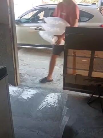 A person in a red shirt and dark shorts is carrying multiple large, clear plastic bags, possibly filled with ice. The person is walking through a doorway into a room or garage where several similar bags are already piled on the floor. The person is in the process of placing the bags they are carrying onto the existing pile. Outside the doorway, a light-colored car is visible. The floor inside appears to be concrete or a similar rough material. The lighting suggests it is daytime.
