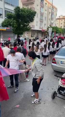 The video shows a crowd of people walking down a street lined with shops, holding a large fabric banner bearing a person's image. Most of the individuals are young adults and teenagers, and some are waving Turkish flags. The banner is predominantly red, white, and blue. The sound of drums can be heard in the background. The scene appears to be a street parade or demonstration. Buildings are visible on either side of the street.