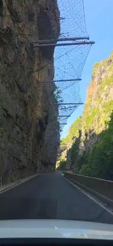 A car drives down a narrow asphalt road between tall rocky cliffs. The cliffs on both sides are covered in wire mesh supported by metal beams, presumably to prevent rockfalls. The mesh has some debris caught in it. The right cliff face has some greenery growing on it. The sky is blue, and the day appears sunny. A road sign is visible on the right.
