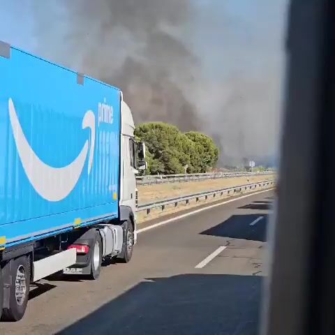 The video shows a bright sunny day with a blue Amazon Prime truck driving on a highway. There is a significant amount of black smoke billowing in the background, indicating a fire. The fire appears to be in a grassy area with some trees. Other vehicles can be seen on the highway, but they are less distinct. The camera is positioned inside another vehicle, likely a passenger car, looking out the side window. The overall impression is that of a serious situation unfolding near the highway.