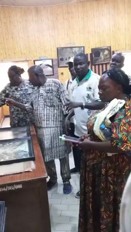 The video depicts a group of people examining framed pictures displayed on a wooden table in what appears to be a gallery or museum. There are six people visible in the video. The woman in the foreground is wearing a colorful patterned dress and holding a cell phone. A man next to her is wearing a white shirt with a green collar and appears to be gesturing towards the pictures on the table. Another man, wearing a striped shirt and glasses, is pointing at a picture in a frame. A woman in a patterned top is standing near him, also looking at the pictures. There are framed pictures on the wall behind them. The room has tan walls and some bamboo paneling. The atmosphere seems to be educational or exploratory, as the group is focused on the displayed pictures.