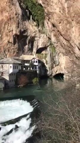 The video shows a building nestled against a large rock cliff, with a river flowing in front of it. There is a small waterfall in the foreground, and the water is clear and turquoise in color. The building is white with a gray roof, and there are a few trees and bushes around it. The rock cliff is mostly bare, with some greenery growing on it. The sky is bright and sunny.