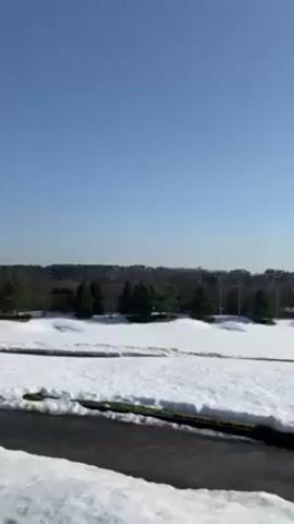 The video begins with a wide shot of a snow-covered landscape under a clear, blue sky. The ground is partially covered in snow, with patches of bare ground and a paved path visible. Trees dot the landscape, and in the distance, buildings can be seen. The camera then pans to the right, showing more of the snow-covered ground, trees, and path. A person wearing a black beanie and a black hooded jacket briefly appears in the frame, looking at the camera. The camera then points straight up at the clear blue sky.