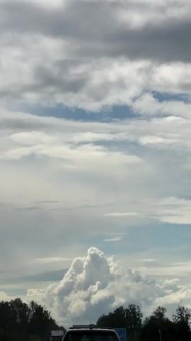 A time-lapse video shows a massive cumulus cloud formation developing in the sky above a highway. The cloud is white and puffy, with a darker base, suggesting it may be a thunderstorm. The sky is partly cloudy with some darker gray clouds scattered around. Trees line the side of the highway, and several vehicles, including a truck and cars, are visible driving on the road. The video appears to be filmed from inside a moving vehicle. The overall impression is one of an approaching storm.