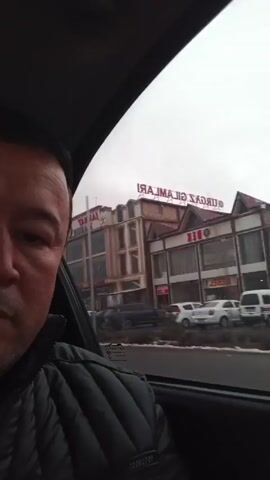 Here is a description of the video:

A man is sitting in a car, looking at the camera. He is wearing a black quilted jacket. The car is moving along a street with buildings on either side. The buildings have signs in a foreign language. It is a cloudy day.