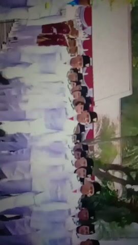 The video shows a group of people in white uniforms with black hats and red accents marching in formation. They are in what appears to be an outdoor setting with trees and a building in the background. The individuals are mostly young men, and some are women. They are marching in a disciplined manner, with their arms extended. Some of the people in the foreground are looking forward, while others are looking to the side. The overall impression is that of a formal procession or ceremony.