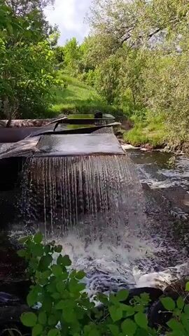 The video shows a narrow, man-made waterfall with water cascading over a metal structure. The surrounding environment is lush and green, with trees and grass on a hillside. The sky appears partly cloudy. The water flow is consistent, creating a foamy splash at the bottom. The scene suggests a natural setting enhanced by human intervention for water flow control.