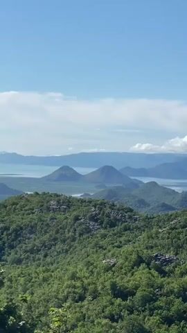 A scenic outdoor shot captures a panoramic view of a vast landscape in Gornja Brova, Montenegro, under a bright, partly cloudy sky. The foreground is dominated by lush green hills densely covered with trees and rocky outcrops. In the midground, a large body of water, likely Lake Skadar, stretches across the scene, flanked by two distinct, somewhat conical hills rising from its shores. Further in the background, a chain of mountains extends towards the horizon, merging with the hazy sky. The camera pans slowly to the right, revealing more of the expansive vista. The sound of a passing vehicle is audible, suggesting the video was filmed from a roadside viewpoint.