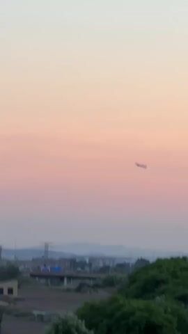 The video shows an airplane flying against a soft pink and orange sky at dusk. The airplane is a silhouette, and it is moving from left to right across the frame. In the background, there are trees, buildings, and power lines. The overall impression is one of peace and tranquility.
