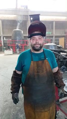 The video shows a man in a welding mask, apron, and protective sleeves in what appears to be an industrial setting. He is standing and looking at the camera with a smile. In the background, there are large industrial objects, possibly metal containers or machinery, some of which are painted red. There are also other people visible in the distance, seemingly working in the same facility. The scene suggests an active workshop or manufacturing environment. The lighting indicates it's daytime. The man is the primary subject and appears to be taking a break or posing for the camera.