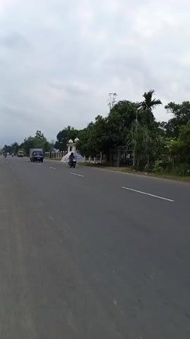 The camera is static, positioned roadside. The scene is a stretch of gray asphalt roadway, divided by white lines. A gray, overcast sky blankets the scene.

On the road, moving from left to right, are various vehicles. First, a black box truck, labeled "POS IND" on its side. Next, a yellow truck travels in the same direction. Motorcycles are also seen, moving in both directions.

To the right of the road is a roadside structure, obscured by a white tarp. Trees and foliage line the right side of the road as well. Some power lines are visible in the distance on the left.