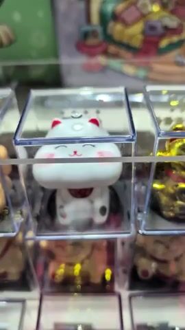 The media shows a close-up of a display case filled with small, individually packaged figurines of Maneki-neko, also known as "beckoning cats," a common Japanese figurine believed to bring good luck. The central figurine is white with red accents on its ears, eyes, and a small collar. It has rosy cheeks and its eyes are closed in a happy expression. The figurines are arranged in rows and columns within clear plastic boxes. The background is blurred, but hints of other colorful displays or packaging are visible, suggesting a retail or market setting. The lighting is bright, indicating it is daytime or well-lit indoors. There is no visible text. The scene evokes a sense of charm and perhaps the anticipation of winning a prize, common in gachapon or claw machine displays. The setting, a display of these popular trinkets, is often found in Asian markets and entertainment venues.