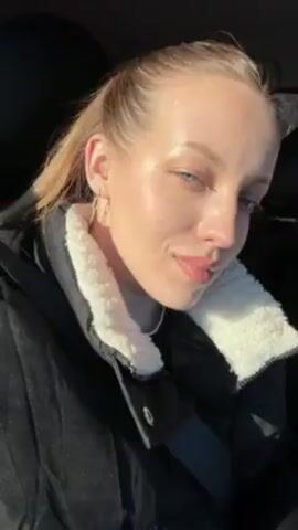 A woman is sitting in the passenger seat of a car. She has fair skin, light hair pulled back, and is wearing a black coat with a white, fluffy collar. She looks at the camera with a slight smile, occasionally closing her eyes slightly and puckering her lips. The car is moving, as indicated by the changing scenery visible through the windows. Sunlight is shining on her face. She raises her hand to her forehead briefly.