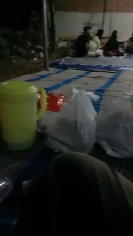 The video captures an outdoor gathering at night. Several people are seated on mats on the ground. In the foreground, there are several white plastic bags, likely containing food. Behind them, a group of approximately six to seven individuals are conversing, some looking towards a laptop placed on the ground. The environment appears to be a backyard or open area, with a brick wall visible in the background and overhead lighting.