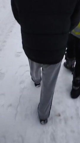Two people are walking through snow in an outdoor setting.  One person is wearing a black jacket with a white patch on the back bearing the number "021", black pants, a black beanie, and black shoes. The other person is wearing a gray and yellow patterned jacket, black pants, a gray beanie, and dark boots. They are walking along a snowy path or road, between snow-covered areas.  Overhead power lines are visible in the background, as are bare trees and some houses in the distance. The sky is overcast.