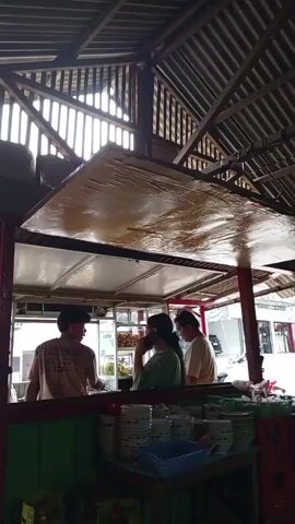 The video shows three people standing behind a counter inside what appears to be a food stall. The counter is made of wood and has shelves displaying plates and bowls. Above the counter is a large, shiny surface, possibly a canopy or roof. The ceiling is made of corrugated metal. The person on the far left is wearing a light-colored t-shirt and is holding a red plastic object. The person in the middle is wearing a green shirt and is holding a phone to their ear. The person on the right is wearing a light-colored t-shirt and is looking down at something on the counter. There are windows in the background that allow light to enter the stall.