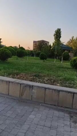 The video shows a park with green grass, manicured bushes, and trees. There is a paved walkway in the foreground and a building in the background. The sky is clear and the lighting suggests it is either sunrise or sunset.