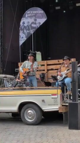 A live music performance is underway outdoors. Two men are playing guitars on the back of a pickup truck that appears to be used as a stage. The truck is light-colored with a yellow trim. The man on the left is wearing a cowboy hat, a patterned shirt, and jeans, and is playing an acoustic guitar. The man on the right is also wearing a cowboy hat, a denim jacket over a light shirt, and jeans, and is playing an electric guitar. Behind them, a drum set is visible. A large black banner with white text and images is displayed behind the performers, advertising "Best of Country Music". The background shows a dark stage area. The ground in front of the truck is paved.