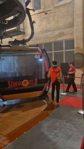 Here's a detailed description of the video:

The video shows a cable car at a station. Initially, the camera focuses on a "SUNWORLD" branded cable car with the number 04. It then pans to another cable car, this one with the number 54 and a logo that is partially obscured. People are inside the second cable car. The camera angle then changes to look upwards at the mechanism above the cable car, highlighting the wheels that facilitate movement along the cable. The wheels appear to be new or in good condition. The environment looks like an indoor station for the cable cars. The lighting is artificial and the overall atmosphere seems industrial due to the visible machinery and construction.