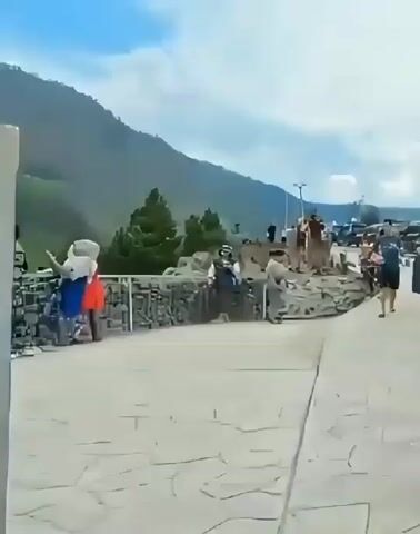 The video shows a group of people standing on a paved area overlooking a valley with rolling green hills and mountains under a cloudy sky. A stone wall with a metal grid is along the edge of the paved area. On the left, two people wearing brightly colored outfits appear to be interacting. Further to the right, several other individuals are standing, some looking out at the scenery. The camera pans slightly to the right, revealing more of the landscape and people. The overall impression is a scenic overlook with visitors enjoying the view.