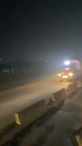 A large truck travels down a road at night. The truck is lit with multiple lights, and the road is lined with concrete barriers. Other vehicles with headlights visible travel in the opposite direction.