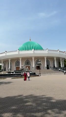 The video presents a wide exterior view of the Amir Timur Museum in Tashkent, Uzbekistan, under a clear blue sky on a sunny day. The prominent, two-story circular building is predominantly white, featuring numerous white columns that support a continuous balustrade along its roofline. Its most striking feature is a large, ribbed turquoise-green dome, topped with a golden finial, sitting centrally on the structure. Arched doorways and windows, adorned with intricate decorative patterns above them, are visible along the building's facade.

In the foreground, a spacious square paved with interlocking bricks stretches towards the viewer. Several multi-tiered fountains are active within rectangular pools positioned directly in front of the museum's main entrances. A few individuals and small groups of people are seen walking across the paved area; one person is riding an electric scooter. Ornate black lampposts are spaced around the square. Green trees line the background, adding foliage to the scene. There is no discernible text visible in the frame.