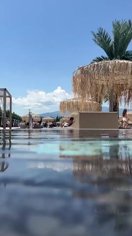 A low-angle shot from inside a swimming pool looking out at a sunny outdoor scene. The water's surface reflects the sky and palm trees. Beyond the pool, people are relaxing on lounge chairs under thatched umbrellas. In the background, mountains are visible under a clear blue sky with some clouds. The scene appears to be a resort or a beach club.
