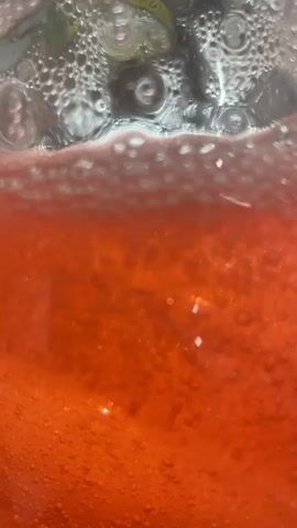 This is a close-up shot of a bubbly, orange-red liquid in a glass. The liquid is carbonated, as evidenced by the numerous small bubbles rising to the surface and forming a frothy layer. The light source illuminates the bubbles, creating a shimmering effect. In the upper portion of the frame, out of focus, appears to be some green foliage, possibly a garnish in the drink, or the background is also out of focus and green. The video is very short and focuses entirely on the texture and movement of the liquid.