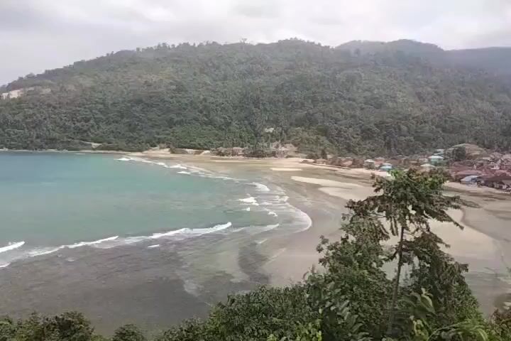 The video shows a wide view of a bay with turquoise water and a sandy beach lined with lush green hills. Waves gently roll onto the shore. In the distance, a few houses and structures are visible along the coastline, nestled at the base of the hills. Trees and foliage are prominent in the foreground, framing the view. The sky is overcast. There are no people or animals visible in the video.