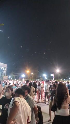 Here's a description of the video:

The video shows a large crowd of people gathered at night in what appears to be an outdoor event or festival. There are several streetlights providing illumination, and some distant buildings are visible in the background. Many of the people in the crowd are standing around, while some are walking. A stage-like structure is visible on the left. There are also numerous small, light-colored objects floating in the air, possibly bubbles. Many people in the foreground appear to be looking towards the center or front of the event. The atmosphere seems lively and social.