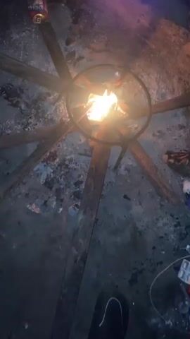 A video shows a close-up, top-down view of a fire burning in a makeshift brazier. The brazier appears to be constructed from a metal ring, possibly a steering wheel rim, supported by wooden beams that radiate outwards from the center. The ground around the brazier is covered in ash and debris, suggesting a temporary or outdoor setting. Various objects are scattered in the background, including what appears to be a bottle of cleaning fluid, a small metal object, and some fabric or clothing. The fire itself is bright and active, with visible flames. The lighting suggests it is nighttime or a dimly lit environment.