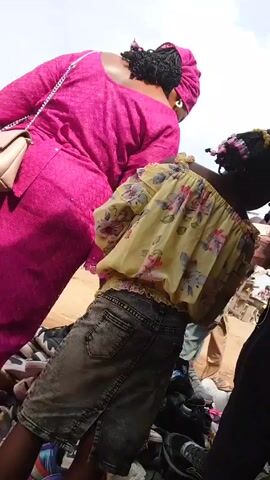 The video shows a woman in a bright pink dress and head wrap standing next to a young girl in a yellow floral top and gray denim skirt. They are both looking at a pile of shoes, which appear to be laid out for sale. A man in black pants stands near the girl, possibly interacting with the shoe selection. The scene appears to be at an outdoor market or roadside sale.  A bicycle passes in the background.