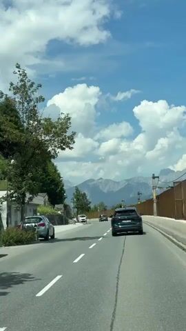 A car is driving on a road with mountains in the background. The road has white dashed lines marking the lanes and a gray shoulder. There are other cars on the road, including a black SUV in front of the camera's car and a white van. On the left side of the road, there is a grassy hill covered in trees. There are small structures with roofs on the hillside, and a wooden fence. The sky is blue with fluffy white clouds.