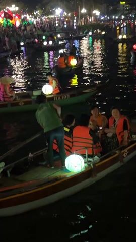 Here is a description of the video:

The video shows a nighttime scene with several small boats on a canal filled with people. Each boat is occupied by passengers wearing life vests and has a lighted orb-shaped lantern attached to the front. A man in a green shirt is rowing one of the boats. The canal is reflecting the lights of the town on the far side of the water. The town has many trees and the boats are filled with people. The overall impression is one of a festive and crowded evening activity.