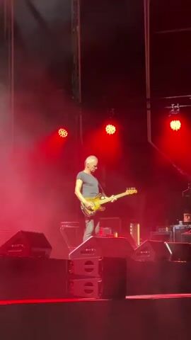 A male musician stands center stage, playing a bass guitar. He wears a dark t-shirt and grey pants. Red stage lights illuminate the scene, casting a hazy glow. Multiple speakers are stacked on either side of the stage. The musician looks down at his instrument as he plays.
