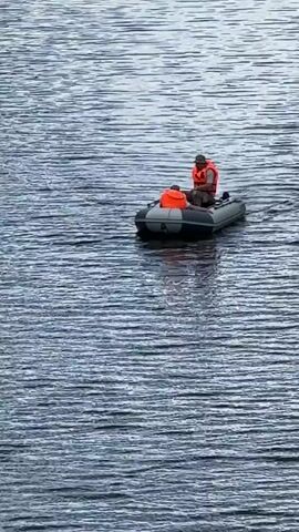 Two people in an inflatable boat are on the water. The person in the back is steering the boat with a motor. Both individuals are wearing orange life vests. The water is choppy with visible ripples.