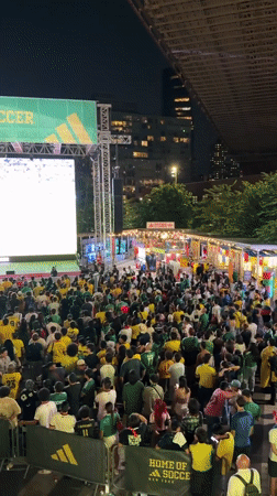 Outdoor crowd watches live soccer under 'Home of Soccer New York' screen