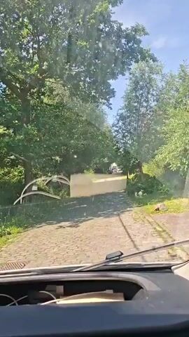 This is a first-person perspective video of a vehicle driving on a cobblestone road lined with trees. The sun is shining, and there are some clouds in the sky. The road appears to be in a rural or suburban area. In the distance, there are some houses and a few other vehicles. The video is shaky, suggesting the vehicle is in motion.