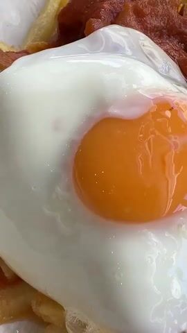 A close-up shot of a plate of food, possibly a breakfast or brunch dish, is presented. The food consists of french fries topped with a rich, red sauce, pieces of meat, and a perfectly fried egg with a bright orange yolk and set white. The egg is the focal point, glistening under the light. The dish is served in a textured bowl, and in the background, a glass of white wine sits on a table covered with a light-colored tablecloth. The lighting suggests an indoor setting, possibly a restaurant. The shot is static with no visible people or interactions.