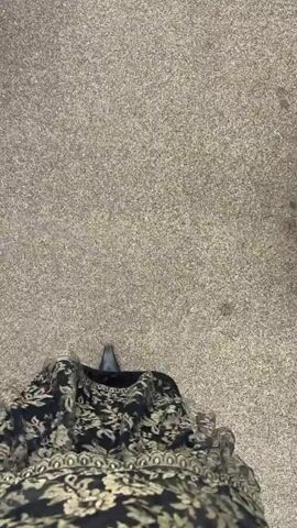 A first-person perspective video shows a person walking on a grey, speckled surface that appears to be pavement. The person is wearing a long, dark-colored skirt with a floral pattern in a lighter shade. Their footwear consists of pointed-toe, black high-heeled shoes with a buckle detail on the strap around the ankle. To the right of the frame, a black object, possibly a piece of clothing or bag, is visible on the ground. The camera angle is directed downwards, focusing on the person's legs and feet as they walk.