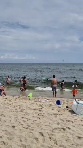 The video shows a crowded beach with people swimming in the ocean and relaxing on the sand. The water is choppy with moderate waves, and the sky is partly cloudy. There are various beach items scattered on the sand, including towels, buckets, and bags. Several people are in the water, some swimming and others standing near the shore. On the beach, people are sitting and standing, some facing the water and others engaged in conversation.