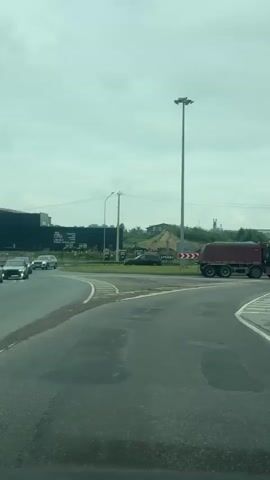 A dump truck turns right at a roundabout. In the background, a tall light pole stands under a cloudy sky. Several cars are visible on the road, and a large, dark building is to the left. The road surface is wet and shows some wear.
