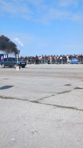 A gray four-door hatchback sits to the left side of the frame in an asphalt field as it spews black smoke out of its exhaust. People stand behind a fence in the background. To the right of the gray car, a second red hatchback burns rubber and kicks up dust as it speeds away from the camera, and toward the crowd of people. A person in black clothing stands to the left of the camera and faces away. The sky is blue and cloudy.