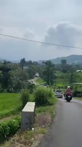 The video shows a slightly elevated view of a road in a rural or semi-rural area. The road is paved and has a white line marking the edge. A white car and a motor scooter with two riders are traveling on the road in the same direction as the camera. Lush green vegetation, including tall grasses, lines the side of the road. In the distance, there are more vehicles and people, and a backdrop of green hills and trees under a cloudy sky. Utility poles with wires run along the side of the road.