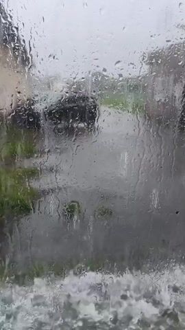 The video shows a view through a rain-streaked window. Outside, a car is visible on a wet road, with greenery along the sides. The rain is heavy, causing water to stream down the windowpane and creating ripples and distorted views of the outside. The bottom of the frame shows a blurry, foamy effect, possibly indicating water splashing or rushing. The overall impression is of a stormy, wet weather scene.