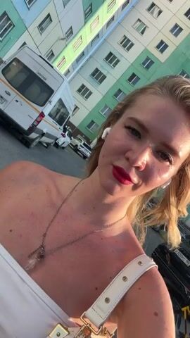 A young woman with blonde hair, red lipstick, and white earbuds is taking a selfie outdoors. She is wearing a white top and a necklace with a crystal pendant. Behind her, a multi-story building with green and beige panels is visible. There are several vehicles in the background, including a white van and a black SUV. Two people on bicycles are also present, one riding a yellow bike. The woman is smiling with her eyes closed.