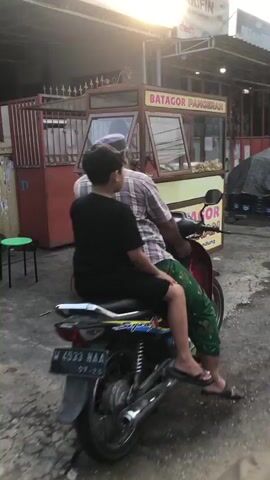 The video captures a rear view of two individuals on a red motorcycle. The person in front, wearing a checkered shirt and a grey head covering, is driving. Behind them, a younger individual in a black t-shirt and green patterned pants is the passenger. Their feet are resting on the motorcycle's footrests, and the passenger's left leg is extended forward. The motorcycle's license plate, reading "M 4533 HAA" and a date "07-25", is visible. The scene appears to be outdoors, possibly in an urban or semi-urban area. In the background, a food stall labeled "BATAGOR PANGERAN" is situated. The stall has glass display cases and appears to be a food vendor. The environment includes a wall, a metal fence, and what looks like tarpaulin sheeting. The lighting suggests it could be late afternoon or early evening, with sunlight casting some warmth on the scene.