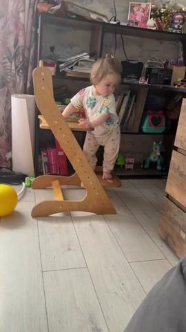 The video shows a toddler standing on a wooden climbing-frame-style chair in a cluttered room. The toddler is wearing a patterned shirt and light-colored pants, with their hair in a small bun. They are reaching up to a bookshelf behind them, which is filled with books, toys, and other items. A yellow ball sits on the floor near the chair. The room has light-colored wood flooring and a patterned curtain. A wooden dresser stands to the right of the bookshelf. A TV hangs on the wall in the background.