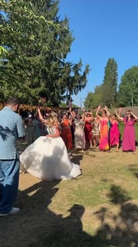 The video shows a bride in a white wedding dress throwing a bouquet backwards to a group of women standing in a semi-circle on a grassy lawn under a clear blue sky. The bride is in the center of the frame, mid-throw, with her back to the camera, her face obscured by her hair. The bouquet is airborne, heading towards the group of women, some of whom have their arms raised in anticipation. The women are dressed in a variety of colorful dresses. In the foreground, a man in a blue patterned shirt and blue pants stands with his back to the camera, facing the bride and the group. Trees and a wooden structure are visible in the background. The lighting suggests it is daytime.